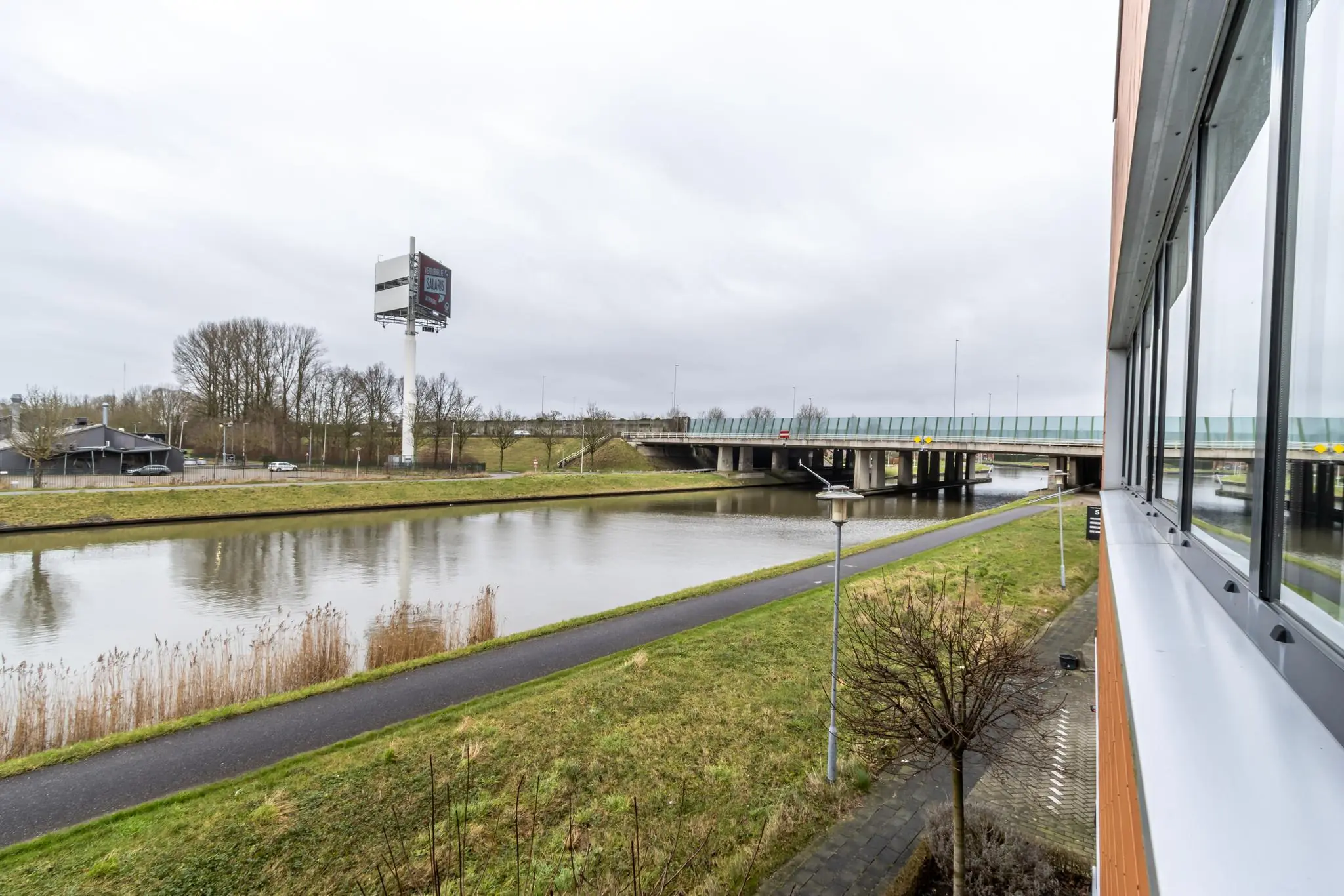 Uitzicht vanaf een gebouw aan de Lage Biezenweg op een kanaal, brug en fietspad onder een grijze lucht.